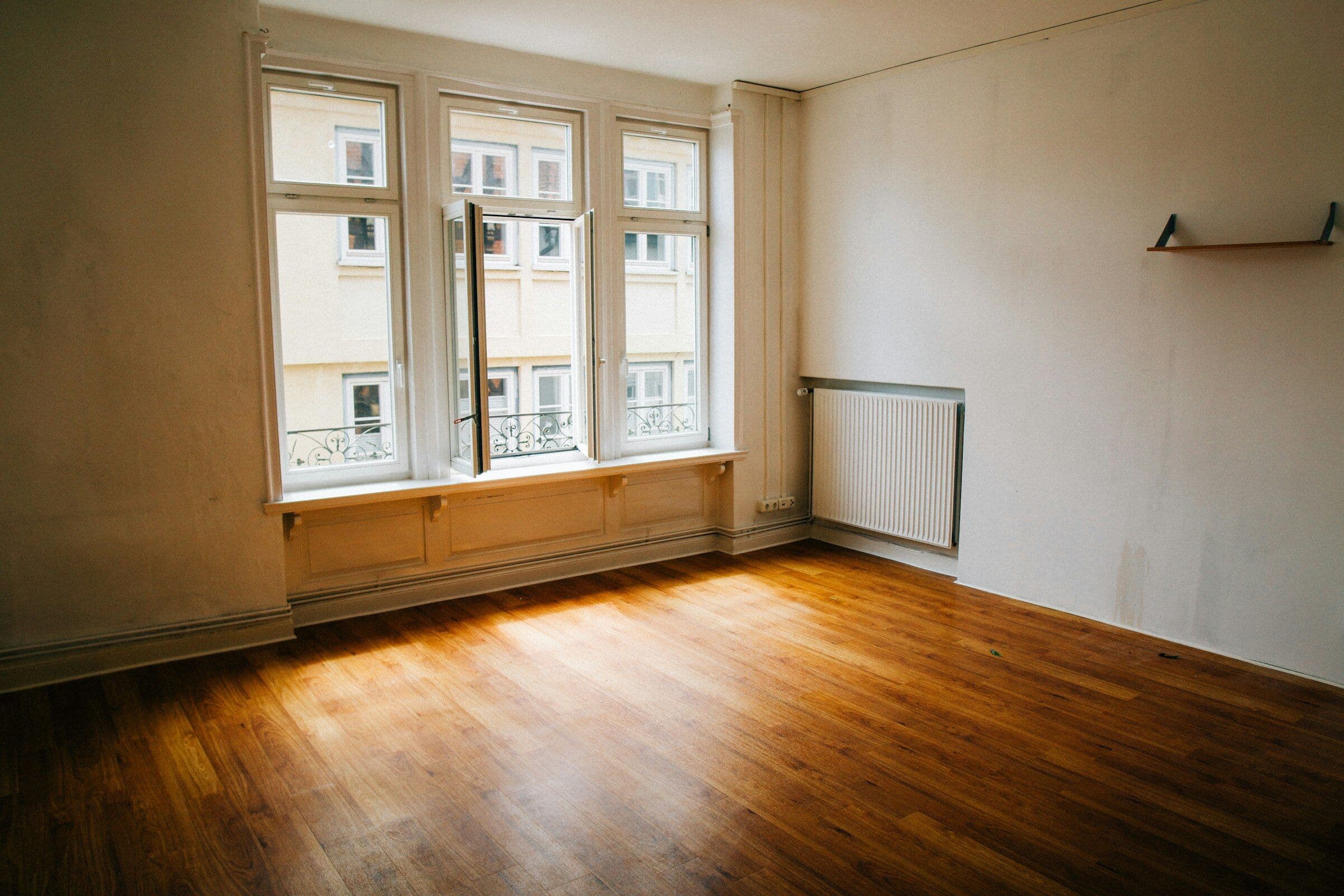 Pièce vide aux murs jaunis, avec un parquet abîmé et un radiateur sous les fenêtres.