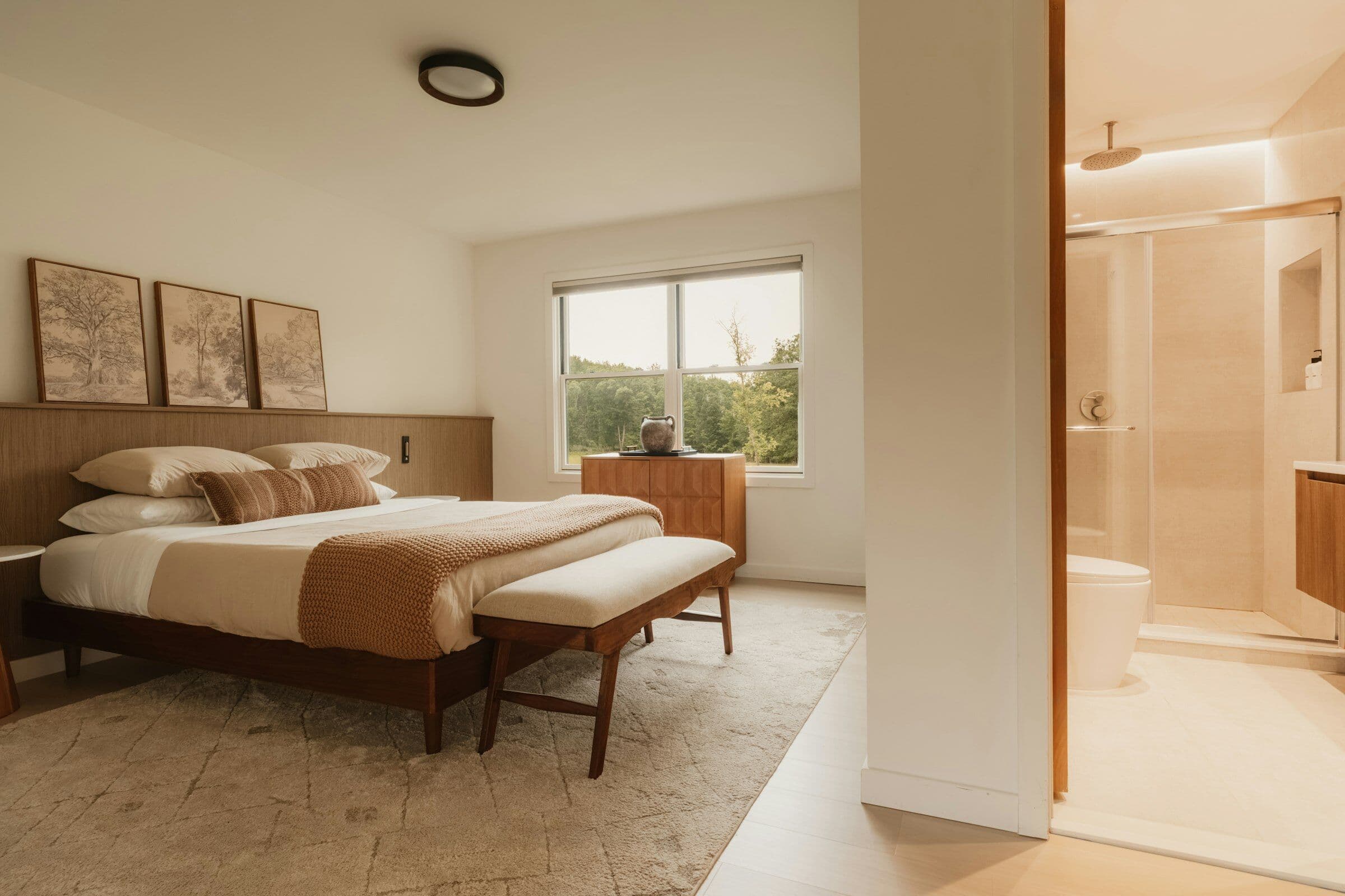 Chambre claire avec un lit en noyer, un banc au pied du lit et une salle d'eau ouverte.
