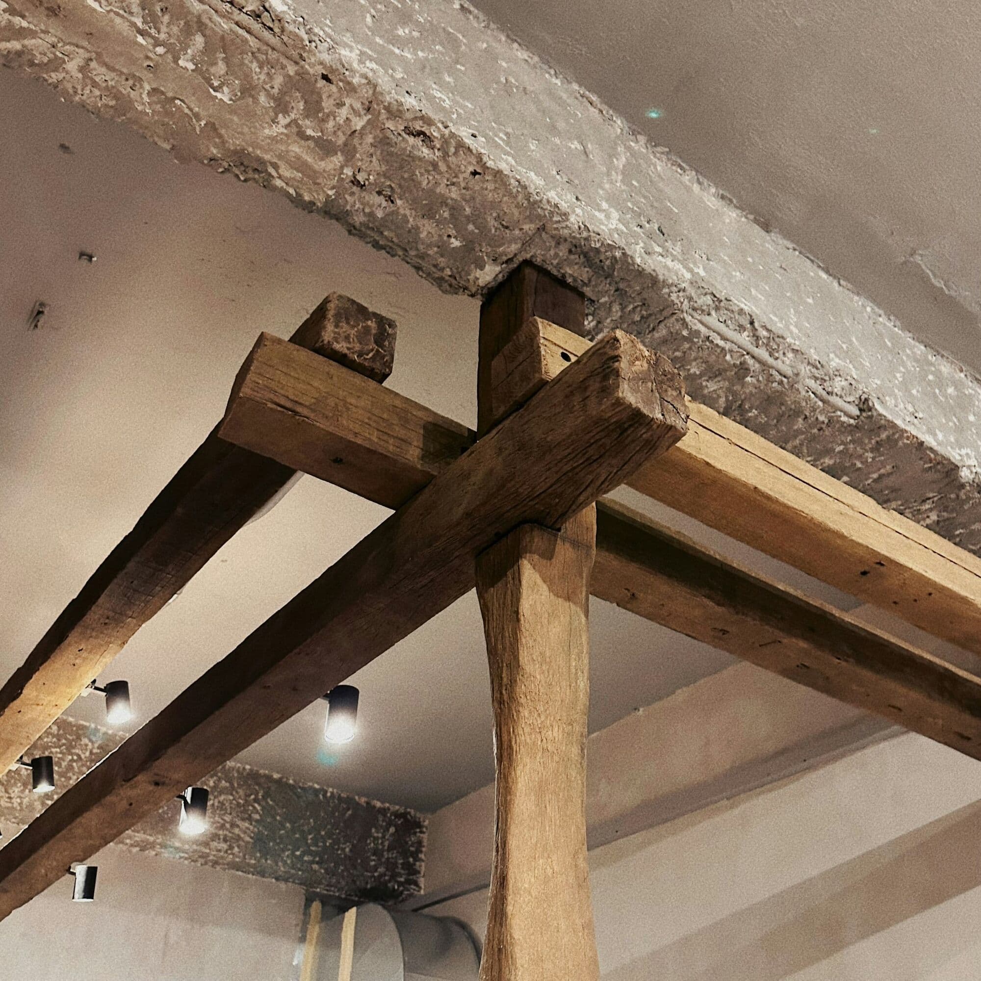 Assemblage de poutres anciennes sous un plafond enduit, éclairé par de petits projecteurs.