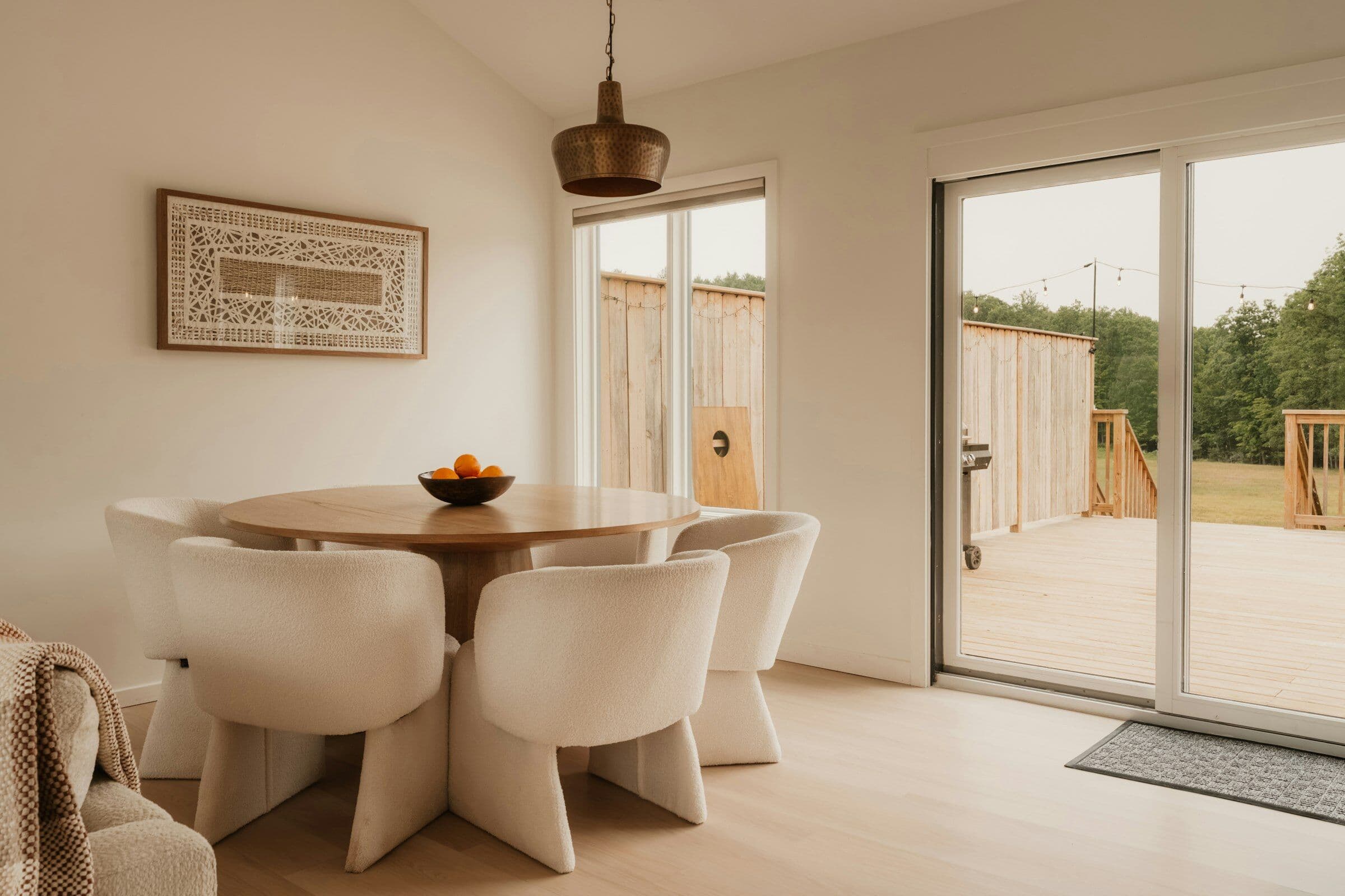 Coin repas baigné de lumière : table ronde en bois, fauteuils blancs et baie ouverte sur une terrasse en bois.