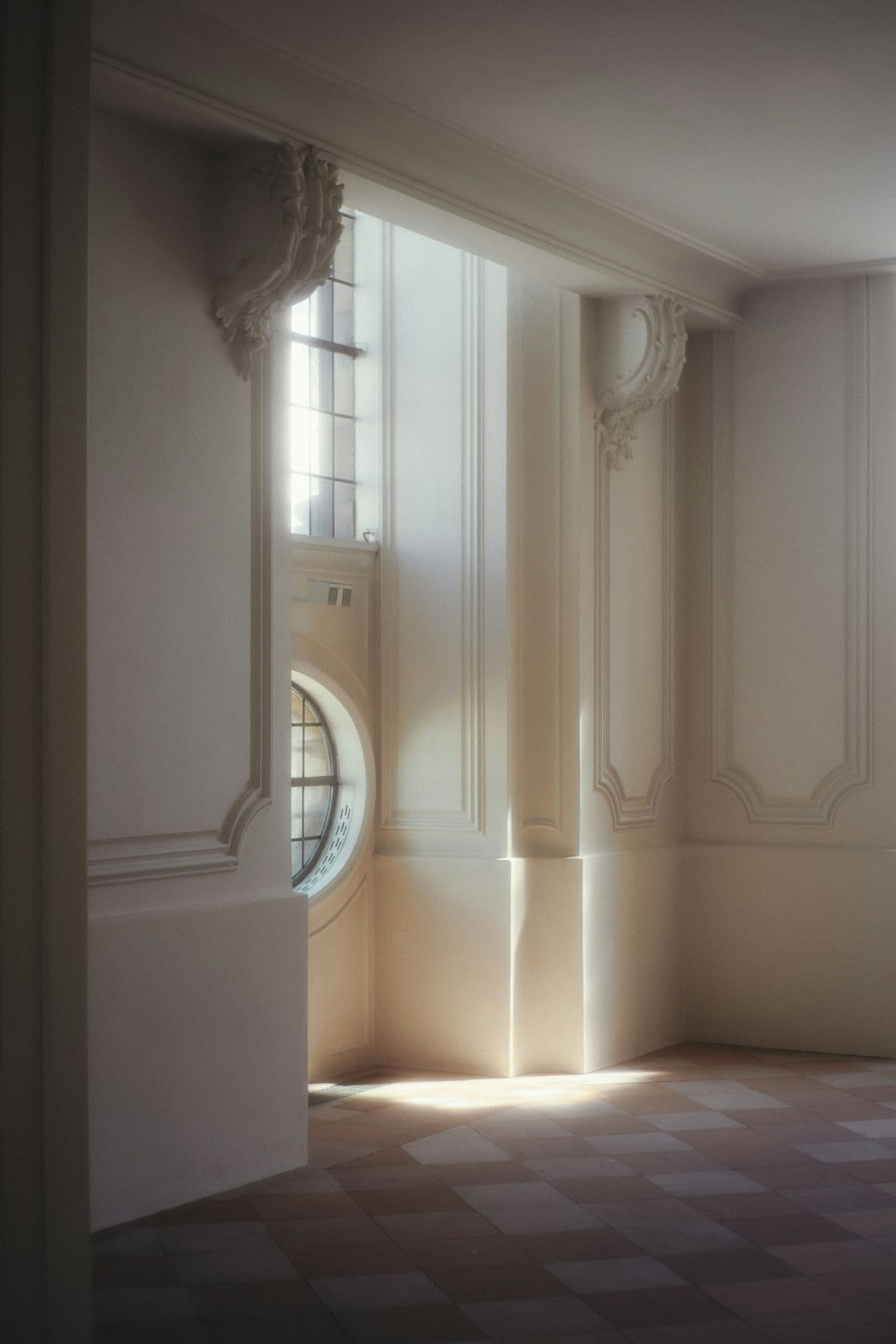 Lumière rasante sur des boiseries et des moulures blanches restaurées, près d'une fenêtre en œil-de-bœuf.