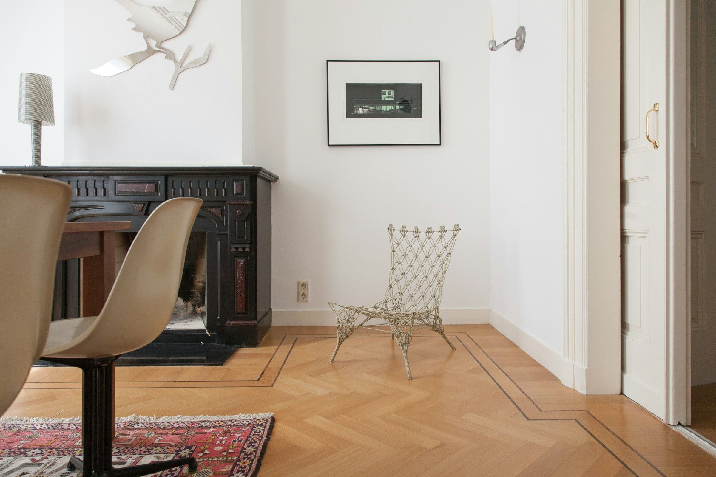 Petit salon blanc au parquet en point de Hongrie, avec une cheminée noire et un fauteuil filaire.