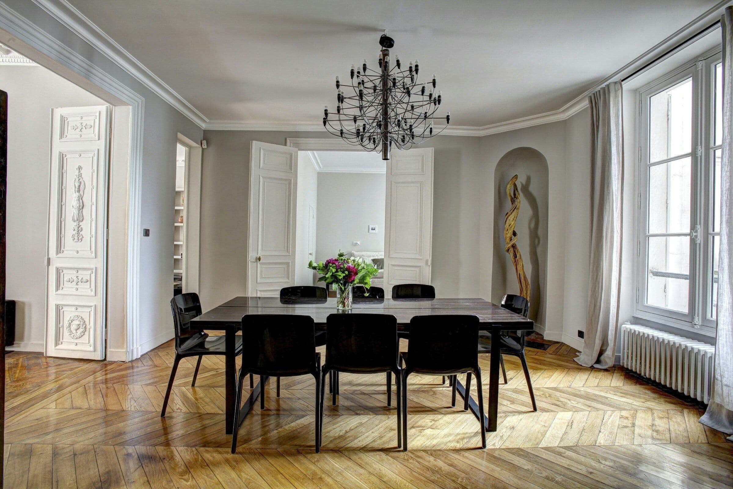 Salle à manger claire avec une grande table noire, un lustre et un parquet en point de Hongrie.