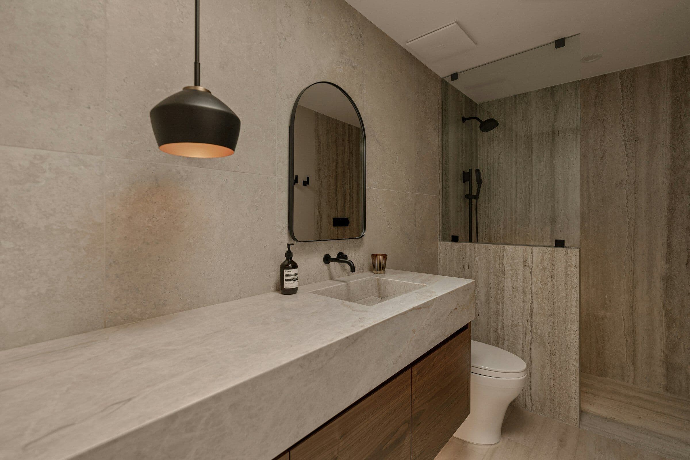 Salle de bain en pierre beige avec un plan vasque long, un miroir arrondi et une suspension noire.