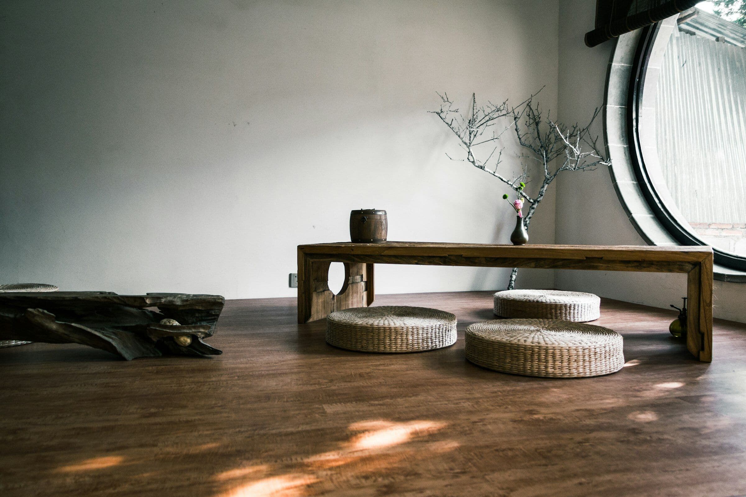Pièce basse au parquet sombre avec une table longue, des coussins de paille et une fenêtre ronde.