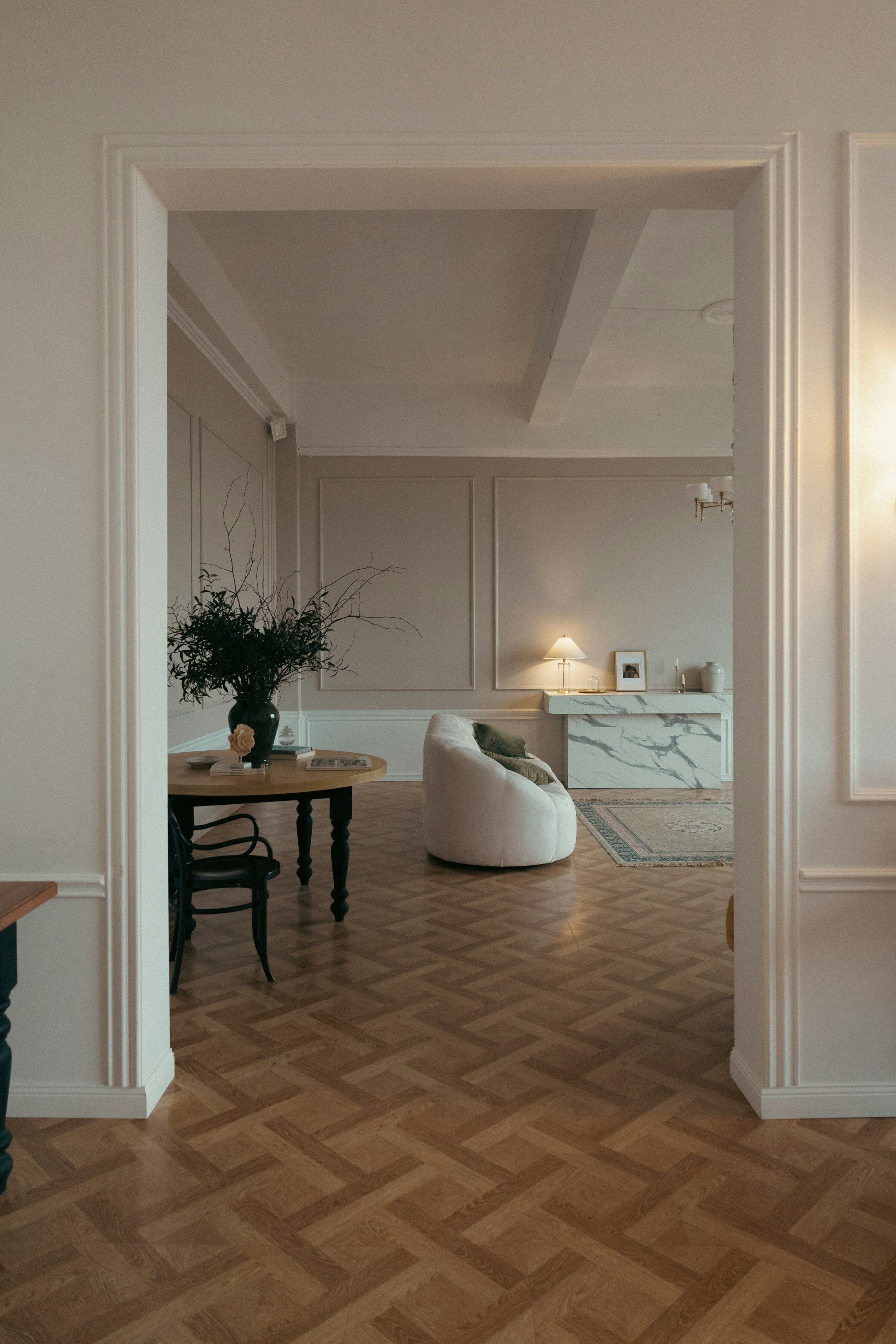 Enfilade de pièces claires vue depuis une porte à moulures : table ronde, fauteuil bouclé et cheminée de marbre.