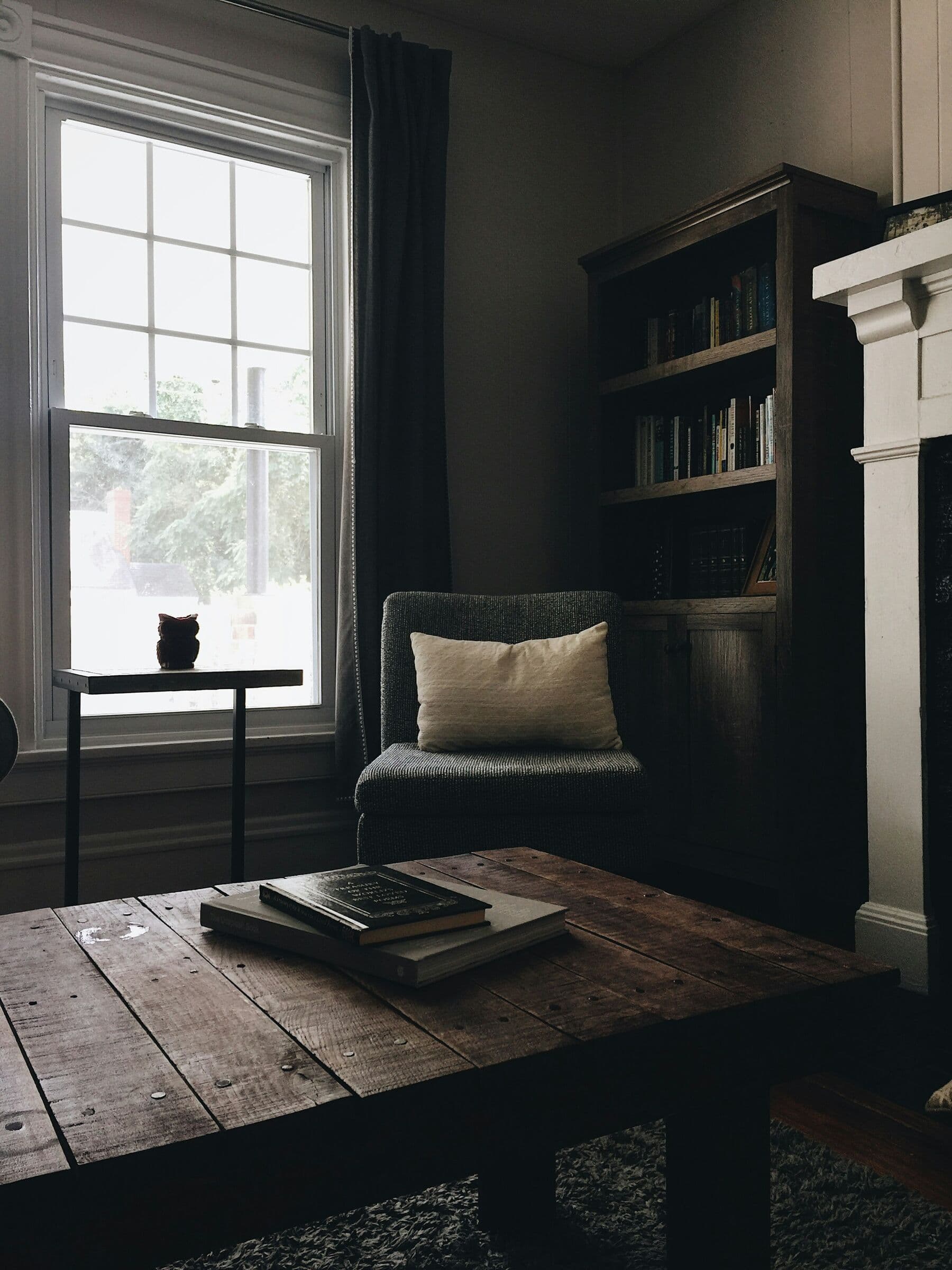 Coin de lecture près d'une fenêtre : fauteuil, bibliothèque sombre et table basse en bois ancien.